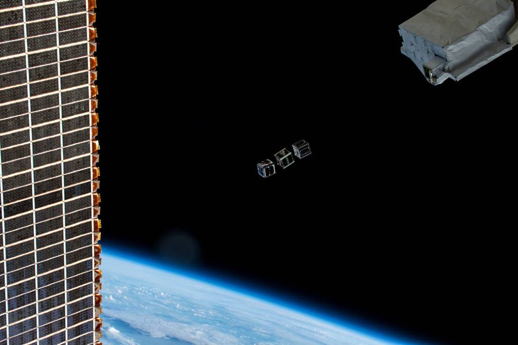 Three small CubeSats drifting away from a deployer outside the International Space Station, with a solar array at left and Earth below